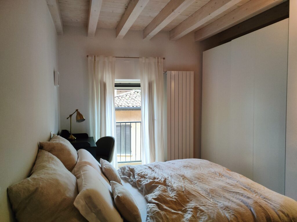 A cozy bedroom in Lakeview Apartment with natural light streaming through sheer curtains, highlighting the rustic wooden beams overhead