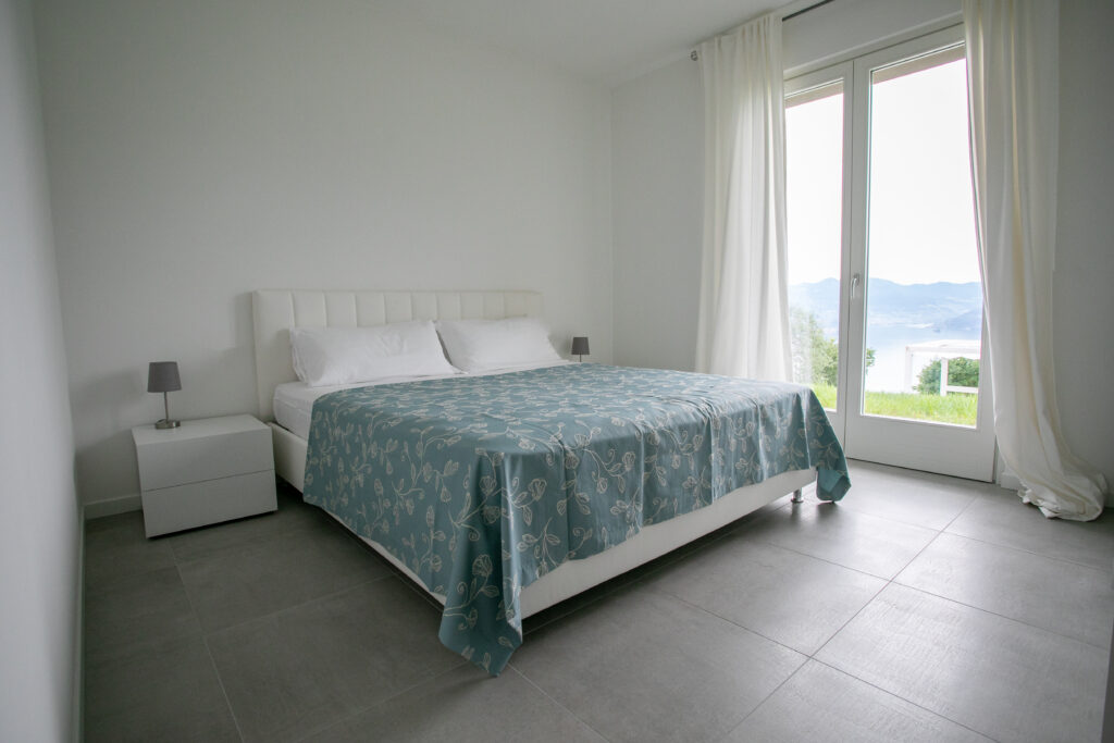 Plush white bed in a luxurious bedroom with floor-to-ceiling windows offering a view of Lake Iseo at Trivia Resort